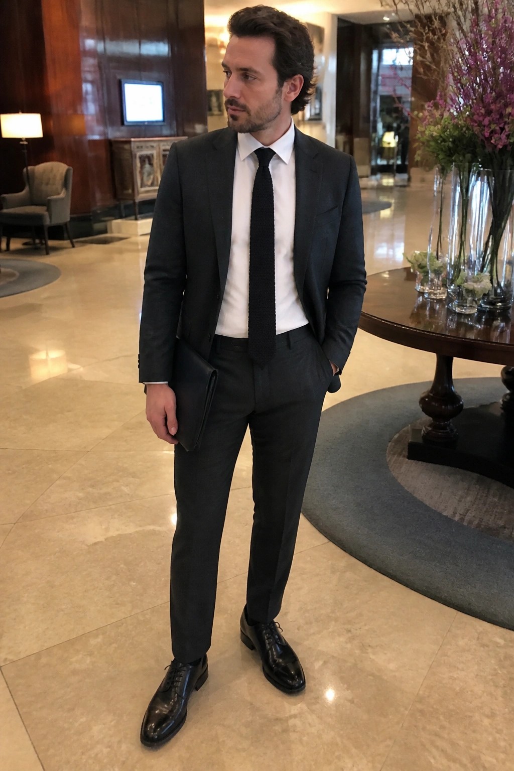 Man in dark suit, white shirt and black tie holding clutch in marble lobby