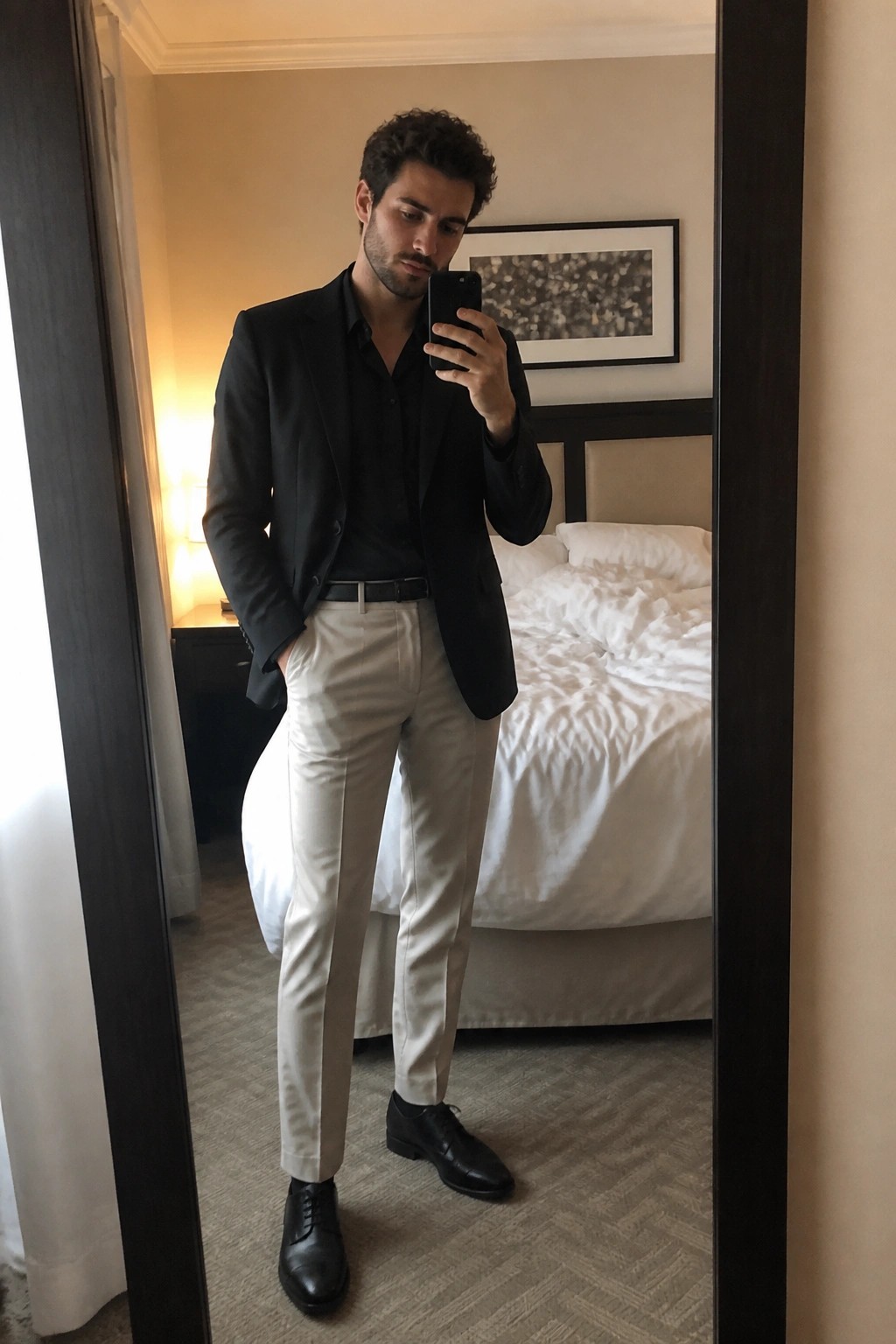 Man in black blazer and beige pants taking mirror selfie in hotel room.