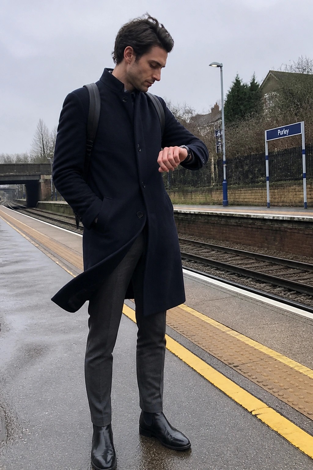 Man in a navy wool overcoat with gray formal pants and black Chelsea boots standing on a train platform.