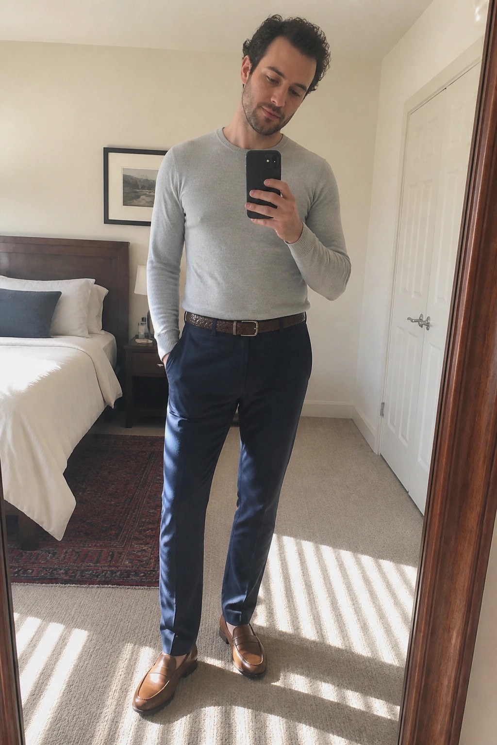 Man in gray sweater and navy pants taking mirror selfie in bedroom