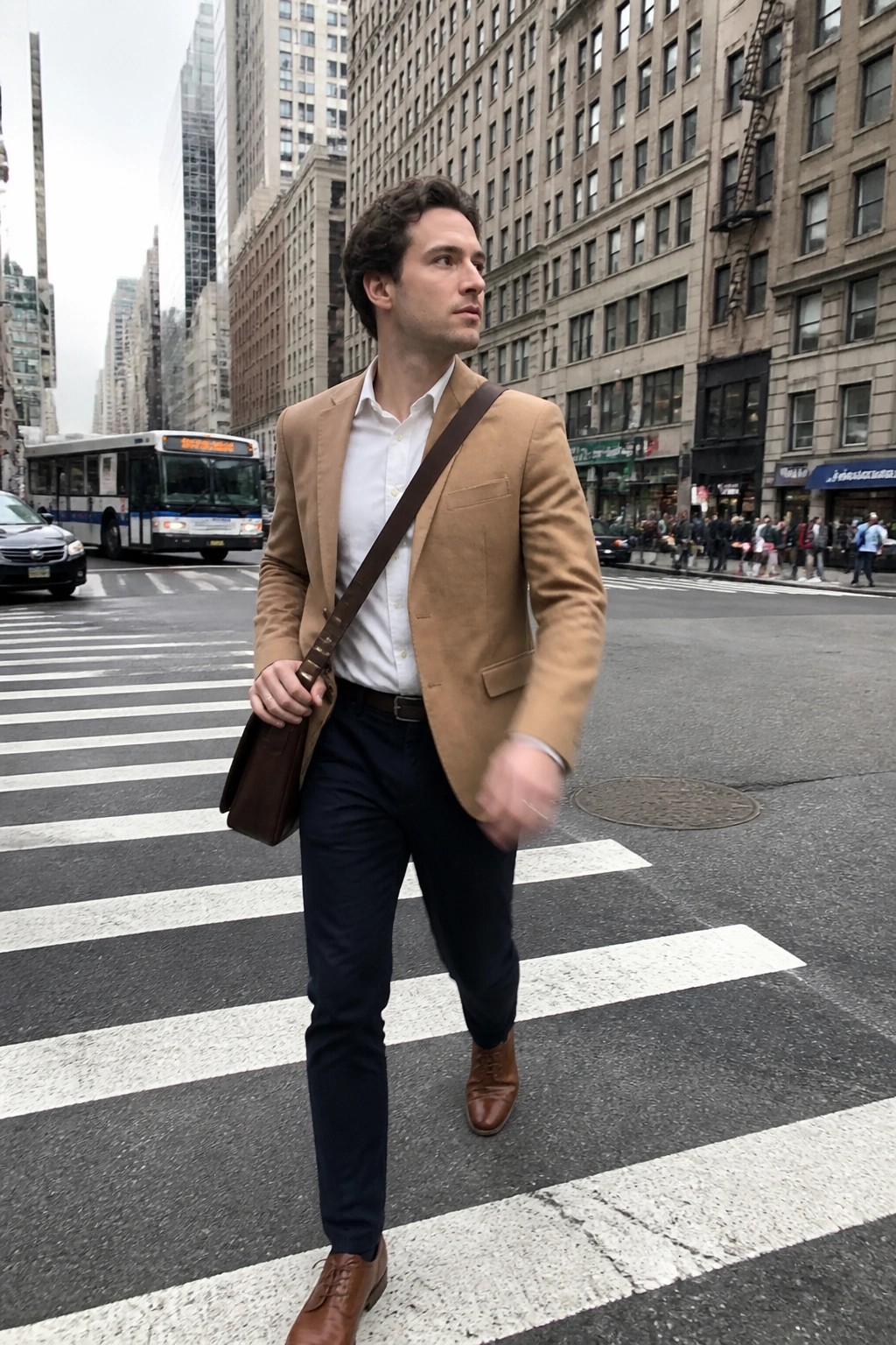 Man in tan blazer crossing city crosswalk with brown leather shoulder bag