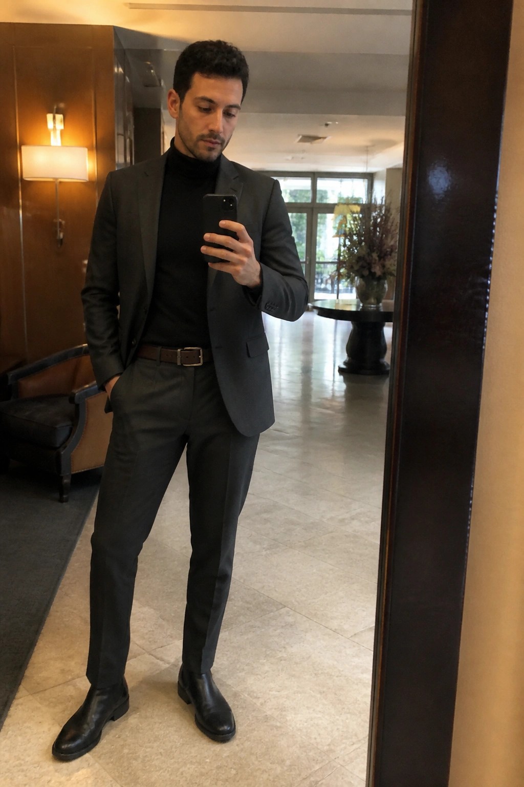 Man in gray suit and black turtleneck takes mirror selfie in elegant lobby.