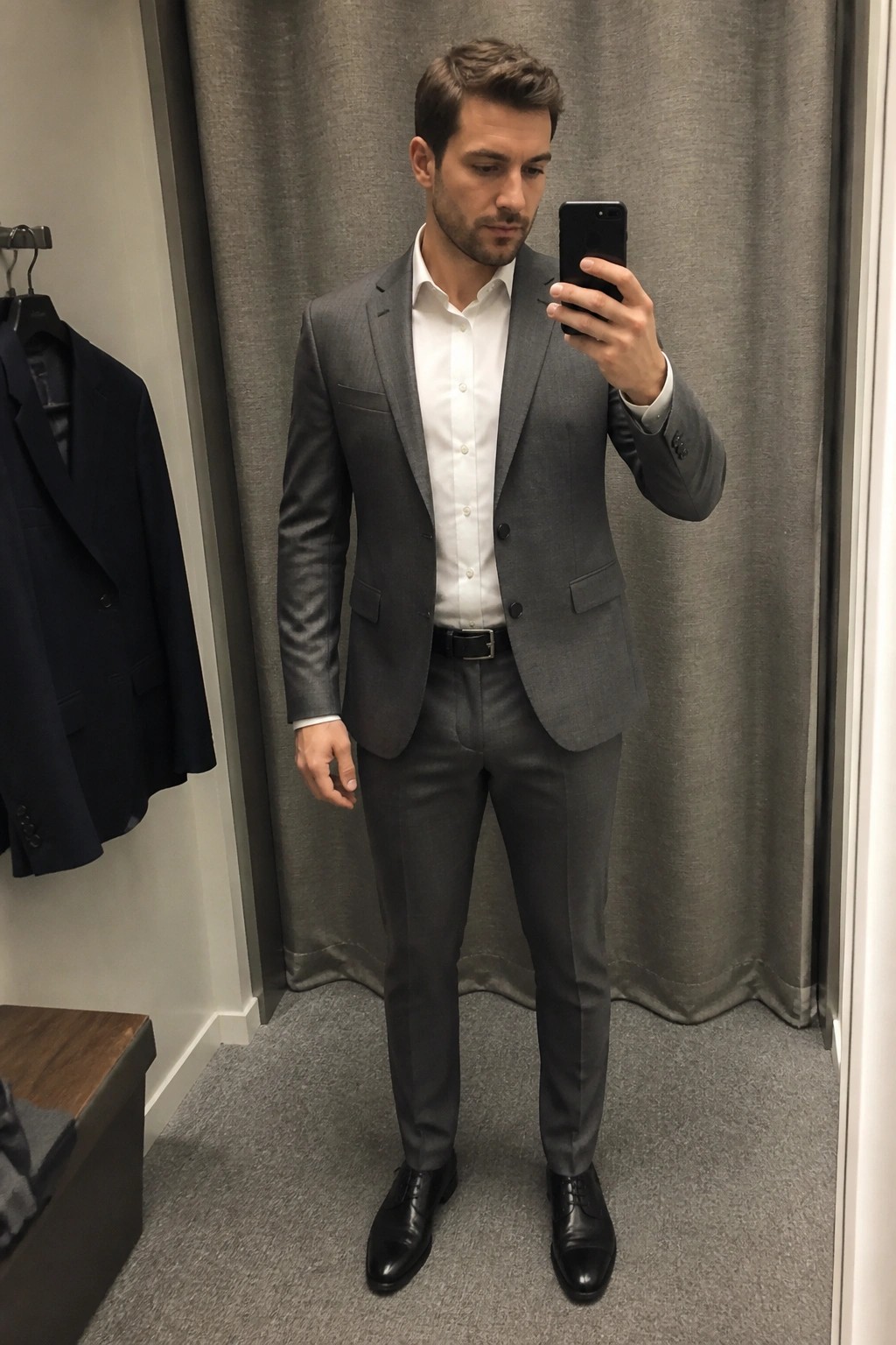Man in gray suit taking mirror selfie with phone in fitting room
