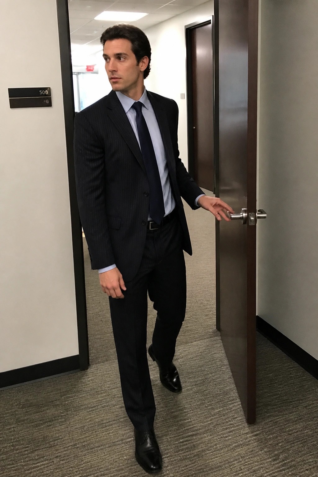 Man in dark pinstripe suit holding open door in office hallway.