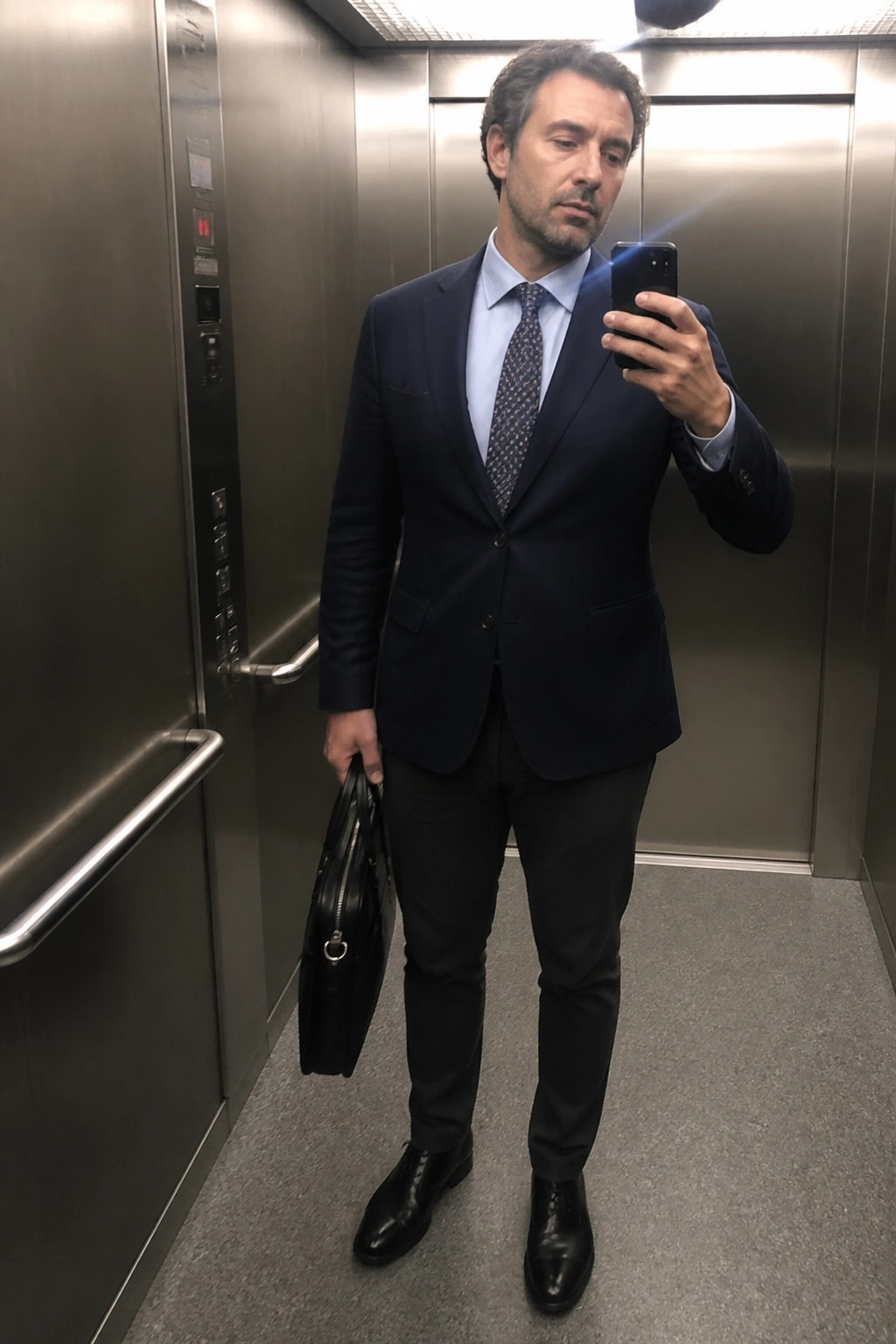 Man in navy suit taking selfie while holding briefcase in elevator