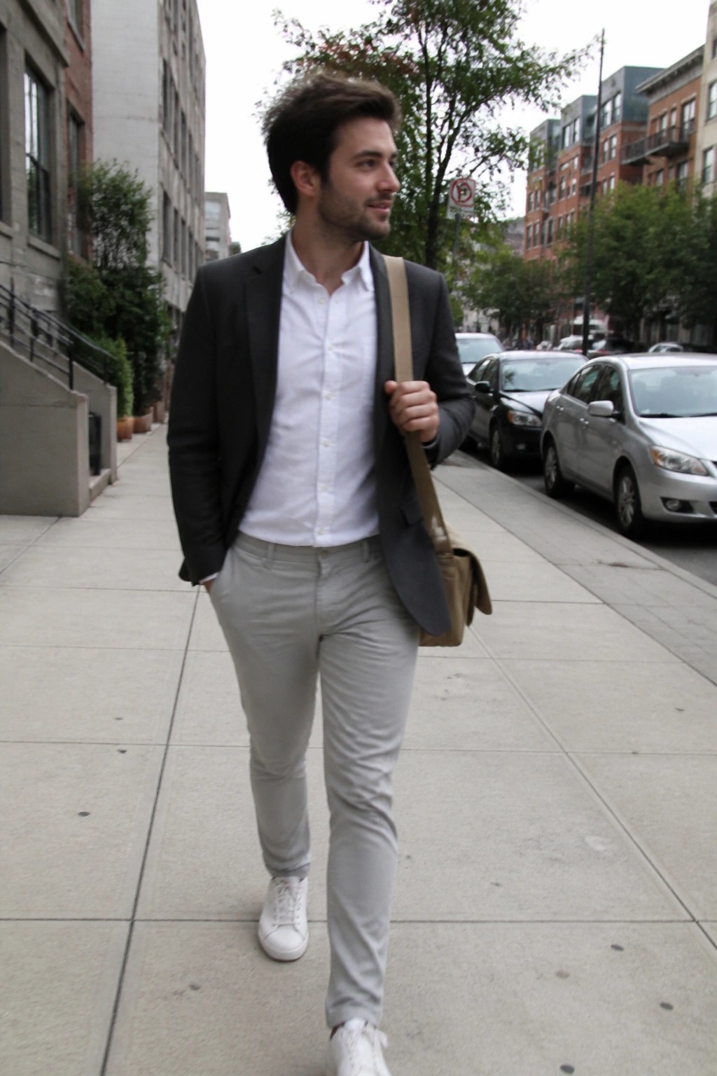 Young man in black blazer and gray pants walks city sidewalk with messenger bag