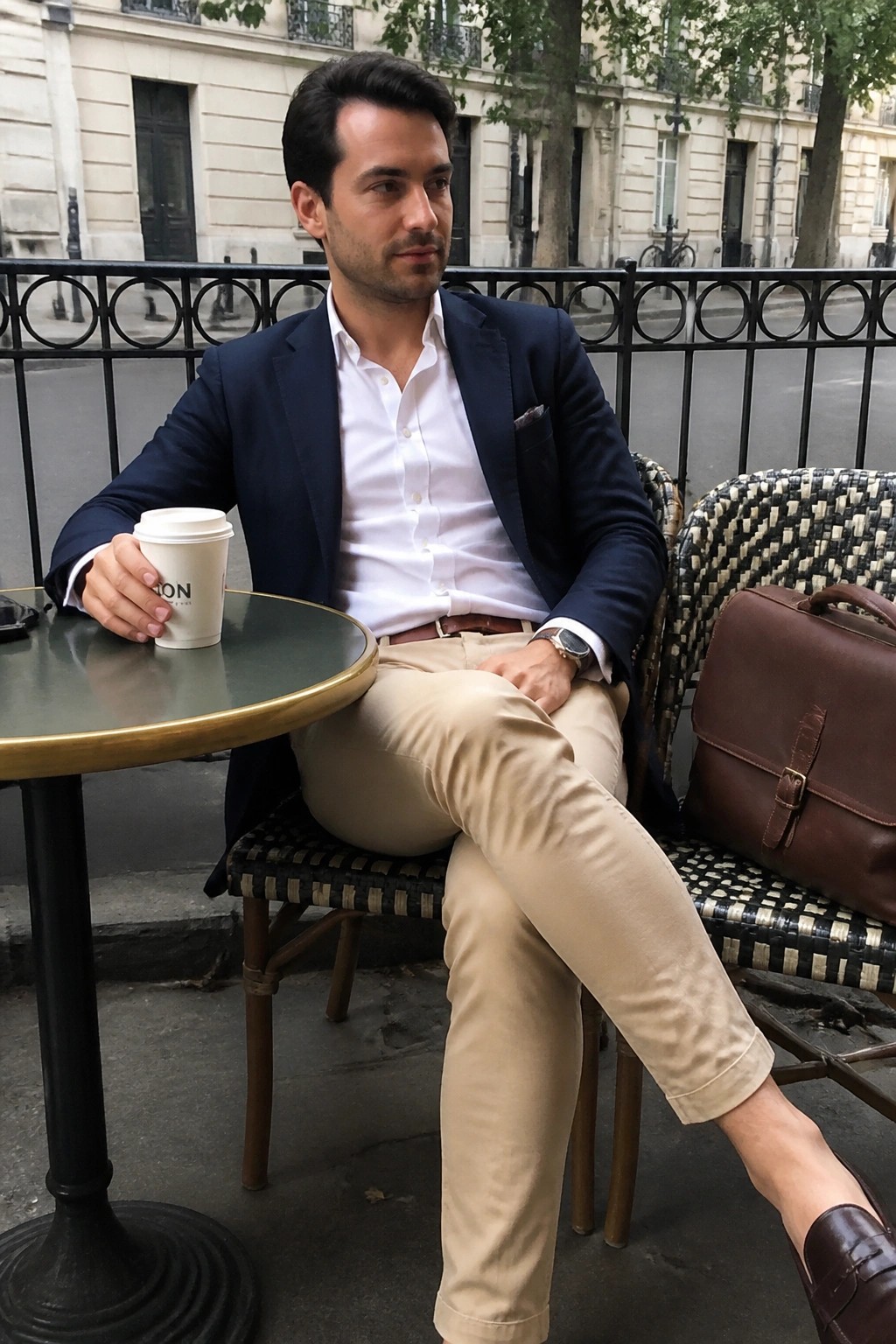 Man in navy blazer and beige pants at outdoor cafe holding coffee beside leather bag