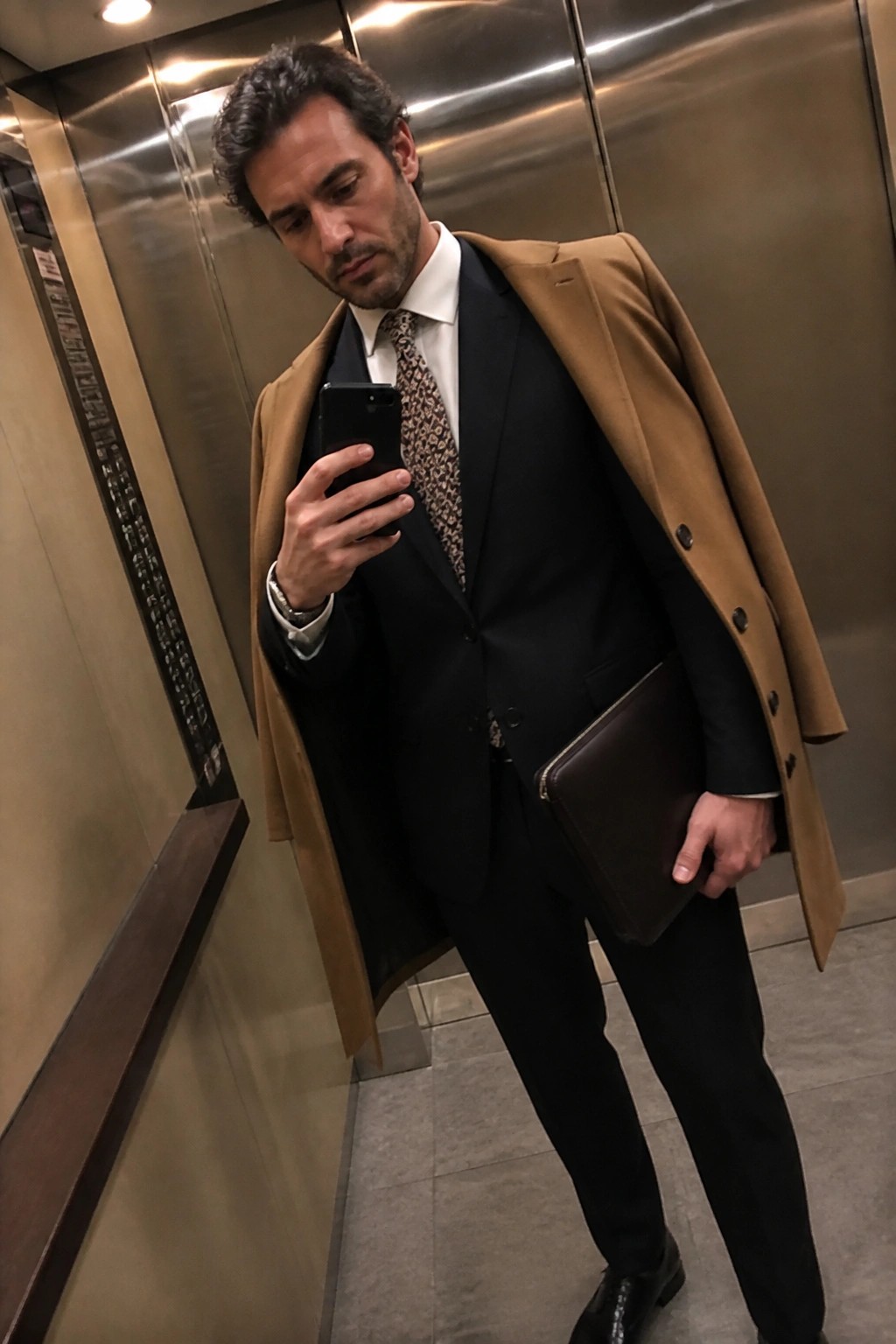 Man in tan overcoat and black suit holding phone and leather portfolio in elevator