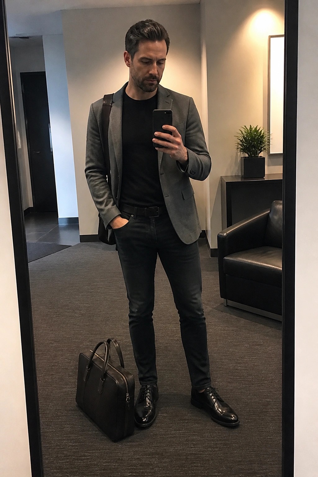 Man in gray blazer taking mirror selfie with phone beside black briefcase
