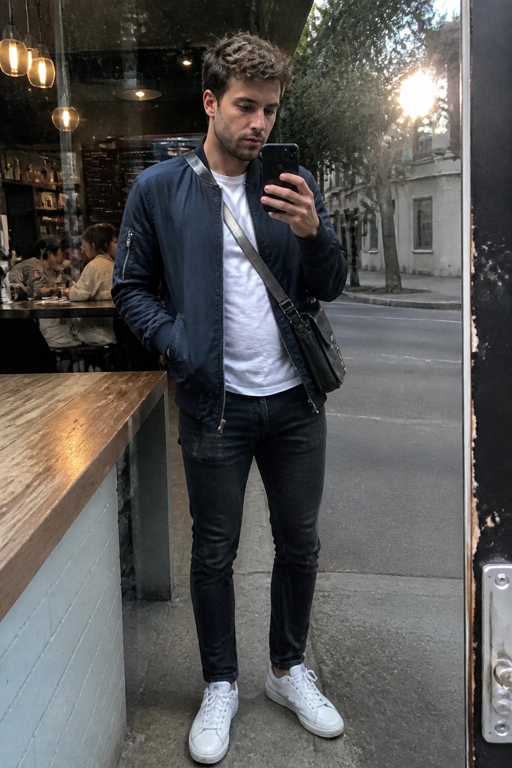 Curly-haired man in navy bomber jacket checks phone outside cafe doorway