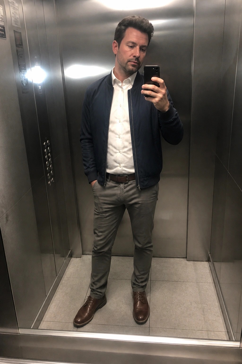 Man in navy bomber jacket taking mirror selfie in stainless steel elevator.