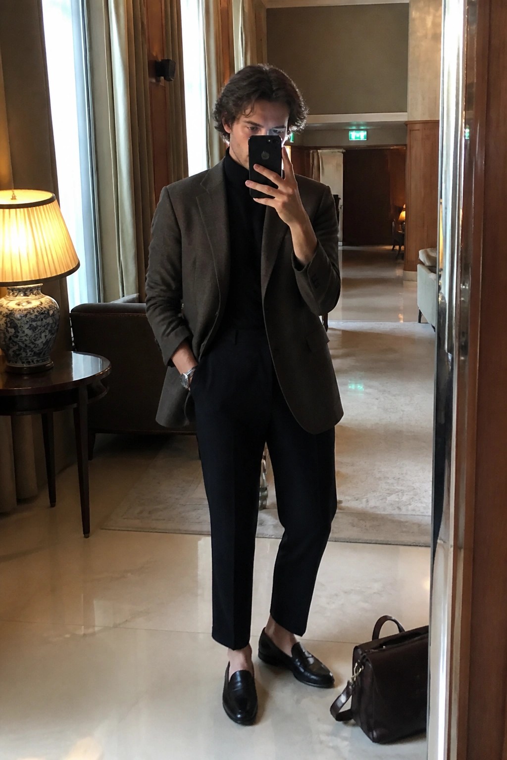 Man in brown blazer over black turtleneck taking mirror selfie in elegant lobby