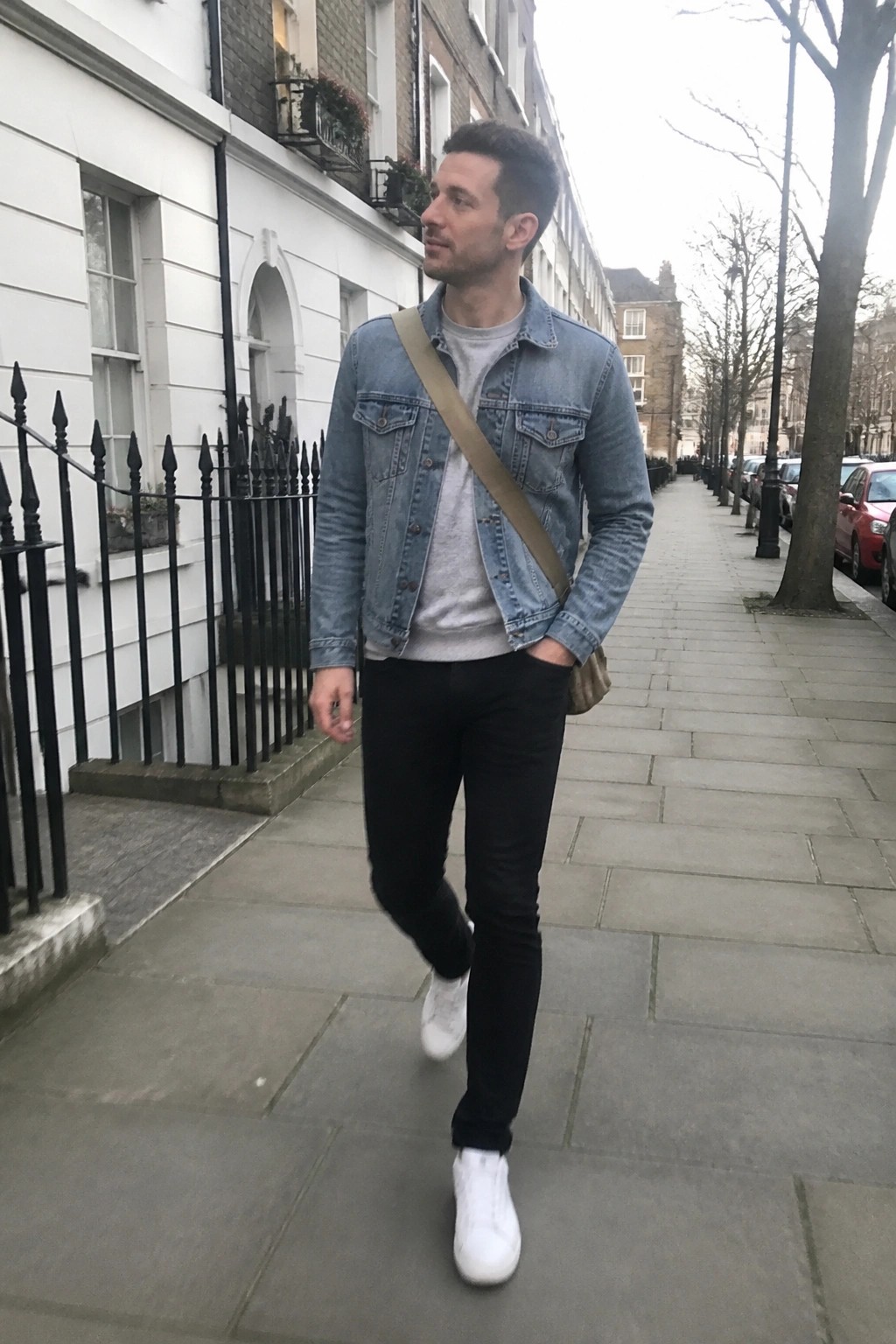 Man in denim jacket and black jeans walking on urban sidewalk with crossbody bag