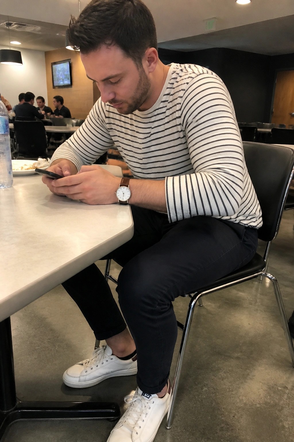 Man in striped shirt and white sneakers checks phone at cafeteria table.