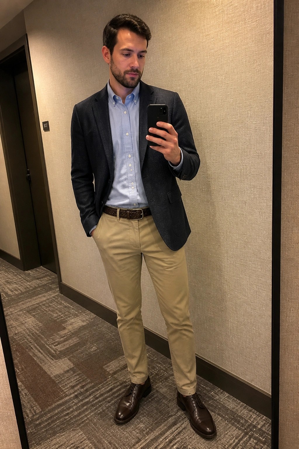 Man in navy blazer, blue shirt, khaki pants taking mirror selfie with phone