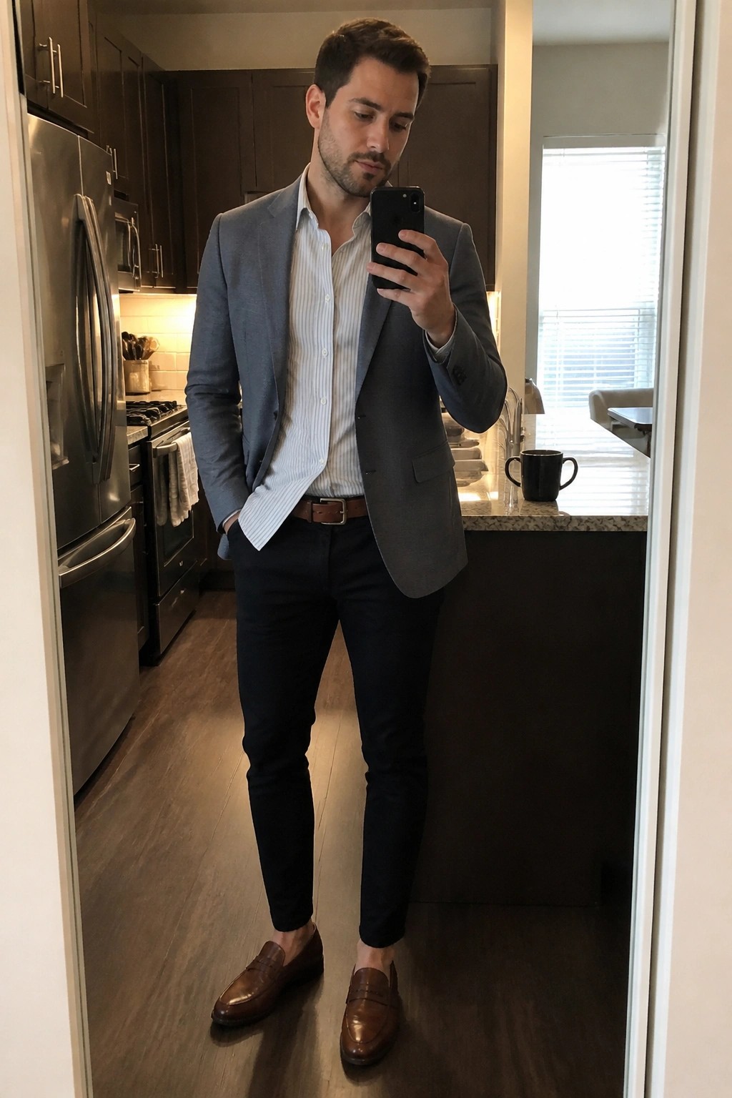 Man in gray blazer over striped shirt holding phone in kitchen mirror