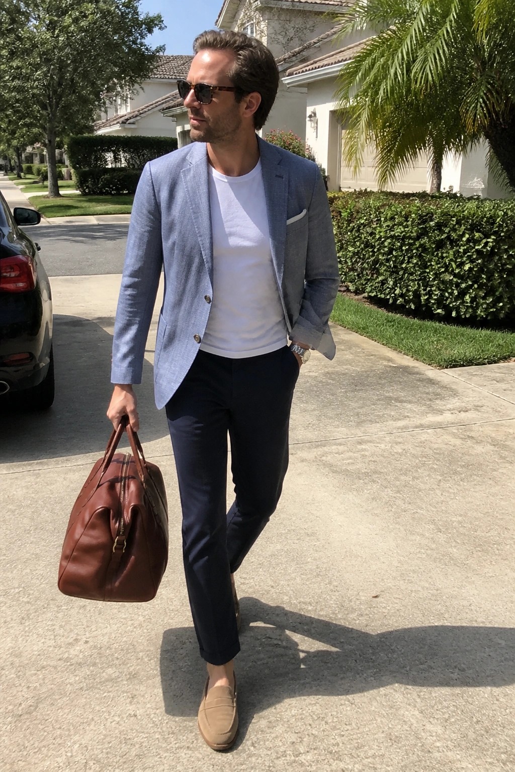 Man wearing light blue blazer over white t-shirt with navy pants, beige suede loafers, and carrying brown leather duffel bag in suburban driveway setting.