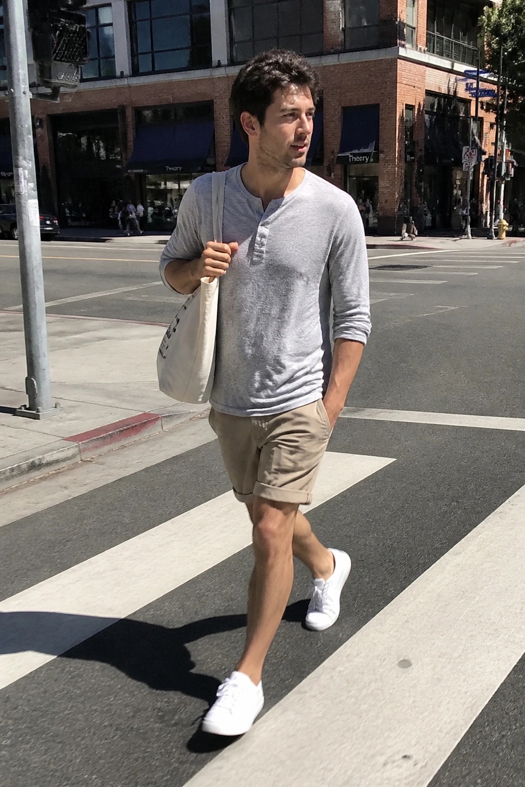 Man in gray henley shirt and khaki shorts crosses sunny urban crosswalk with tote bag