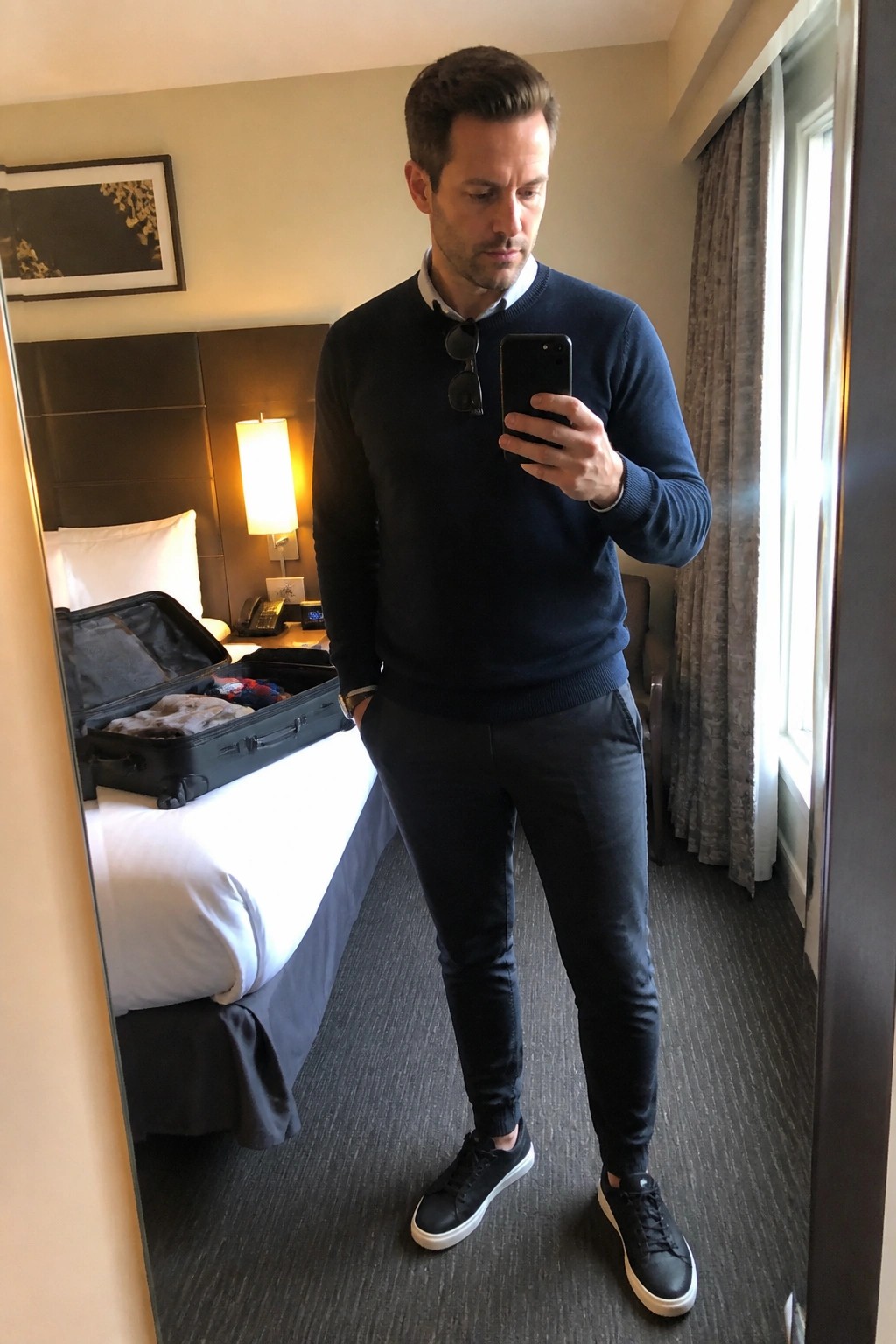 Man in navy blue sweater over light blue collared shirt with dark slim pants and black sneakers with white soles taking a mirror selfie in a hotel room with open suitcase visible on the bed.