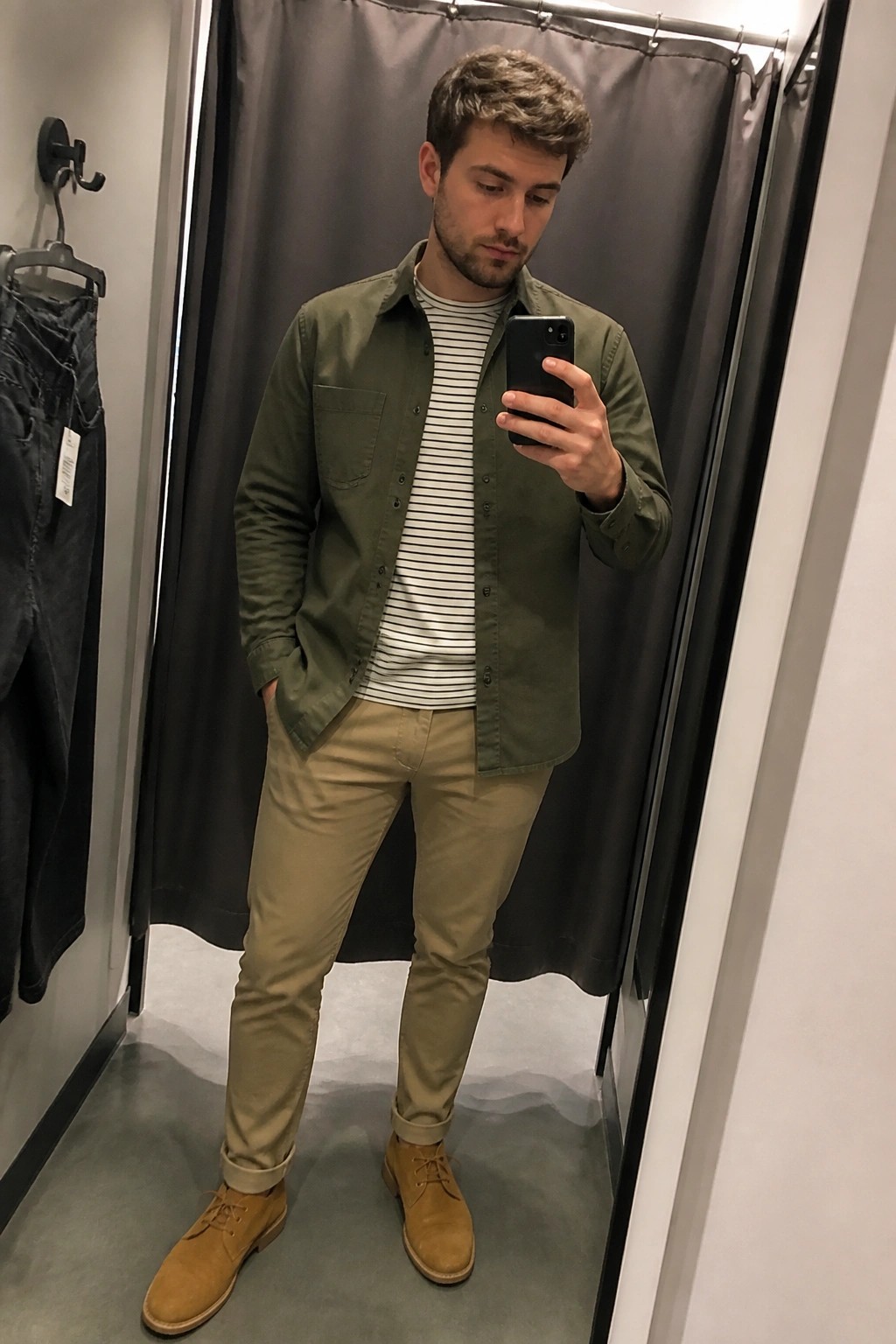 Man in green shirt over striped tee with khaki pants taking mirror selfie