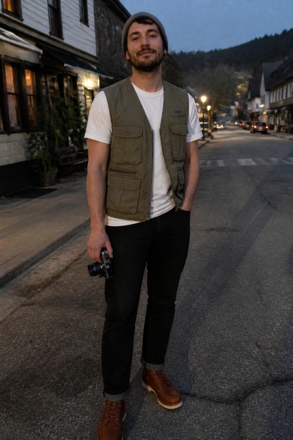 Man in green vest and beanie holds camera on dusk-lit small-town street
