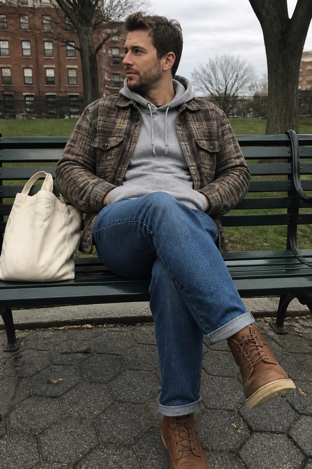 Man in plaid shirt over grey hoodie sits on green bench beside white tote bag