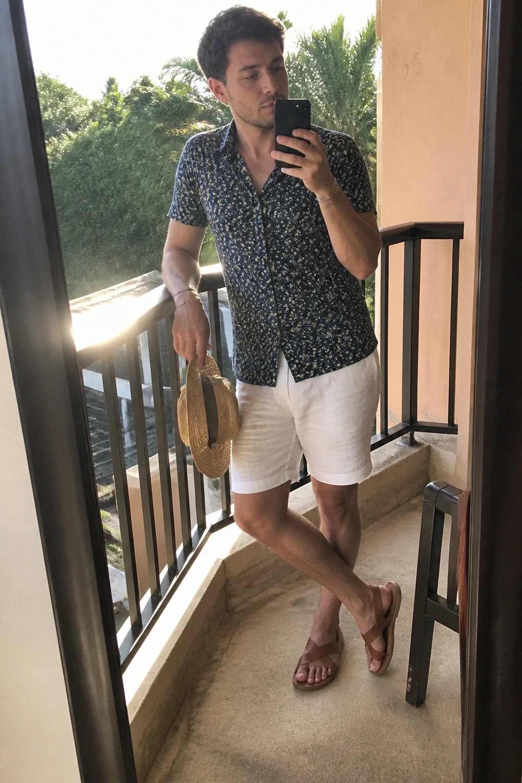 Man on sunny balcony taking selfie in floral shirt, white shorts, holding straw hat