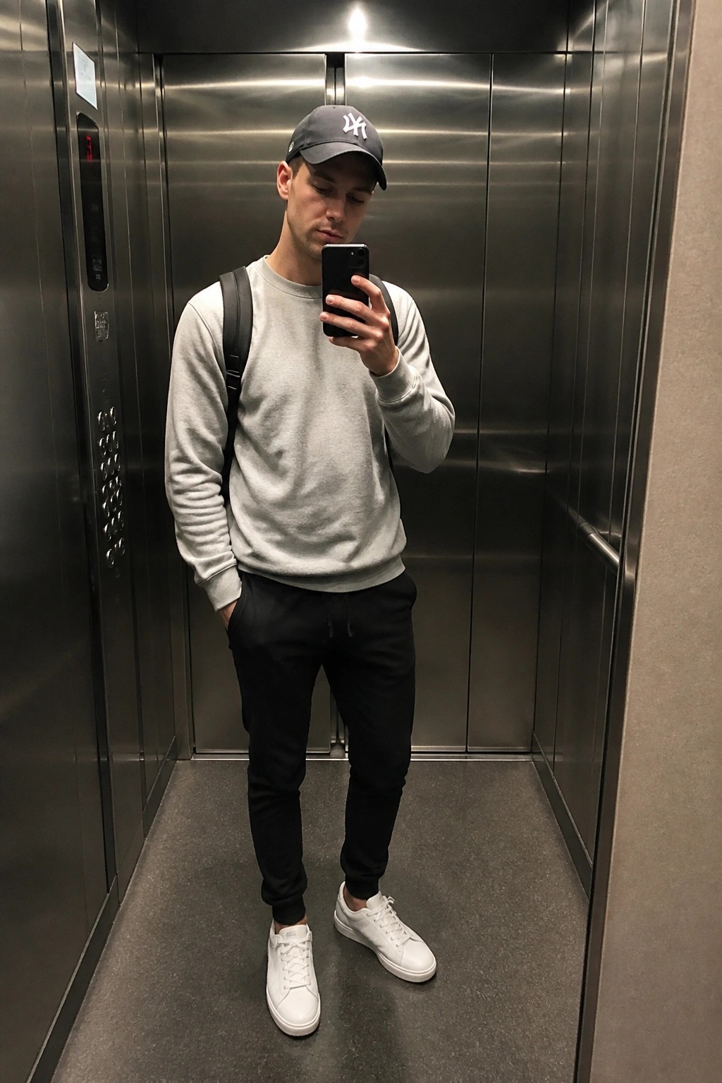 Man in elevator wearing gray sweatshirt, black joggers, white sneakers, black cap, and backpack while holding phone for mirror selfie.