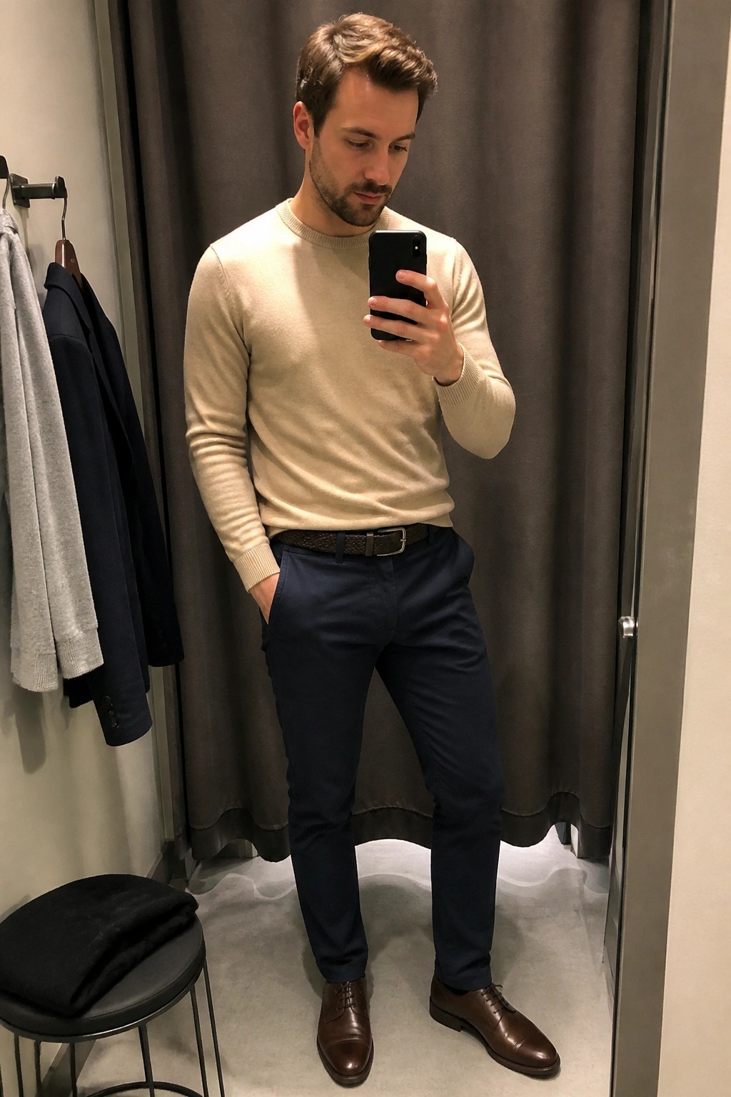 Man in beige sweater and navy pants taking mirror selfie in fitting room
