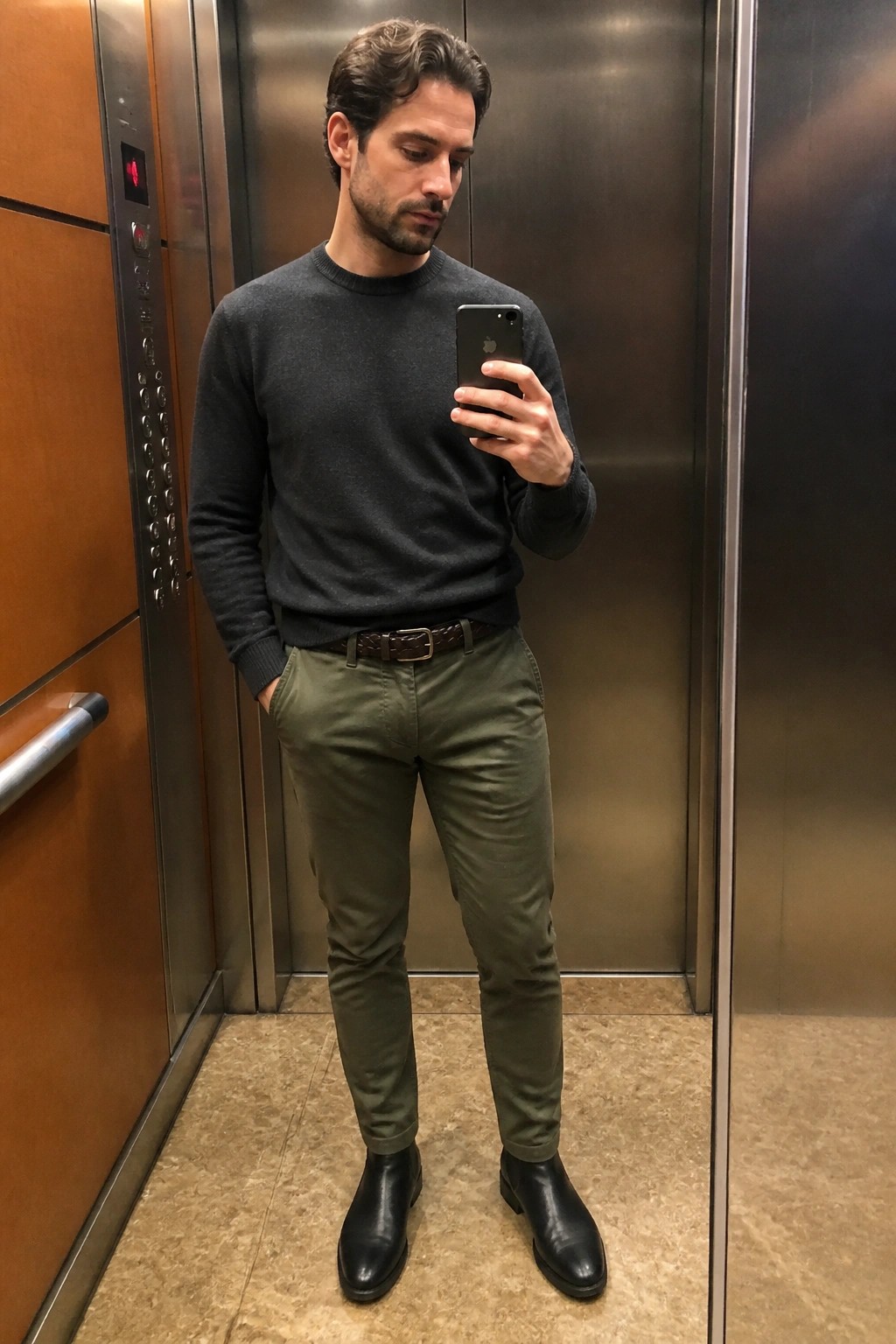Man in gray sweater and olive pants taking mirror selfie in elevator