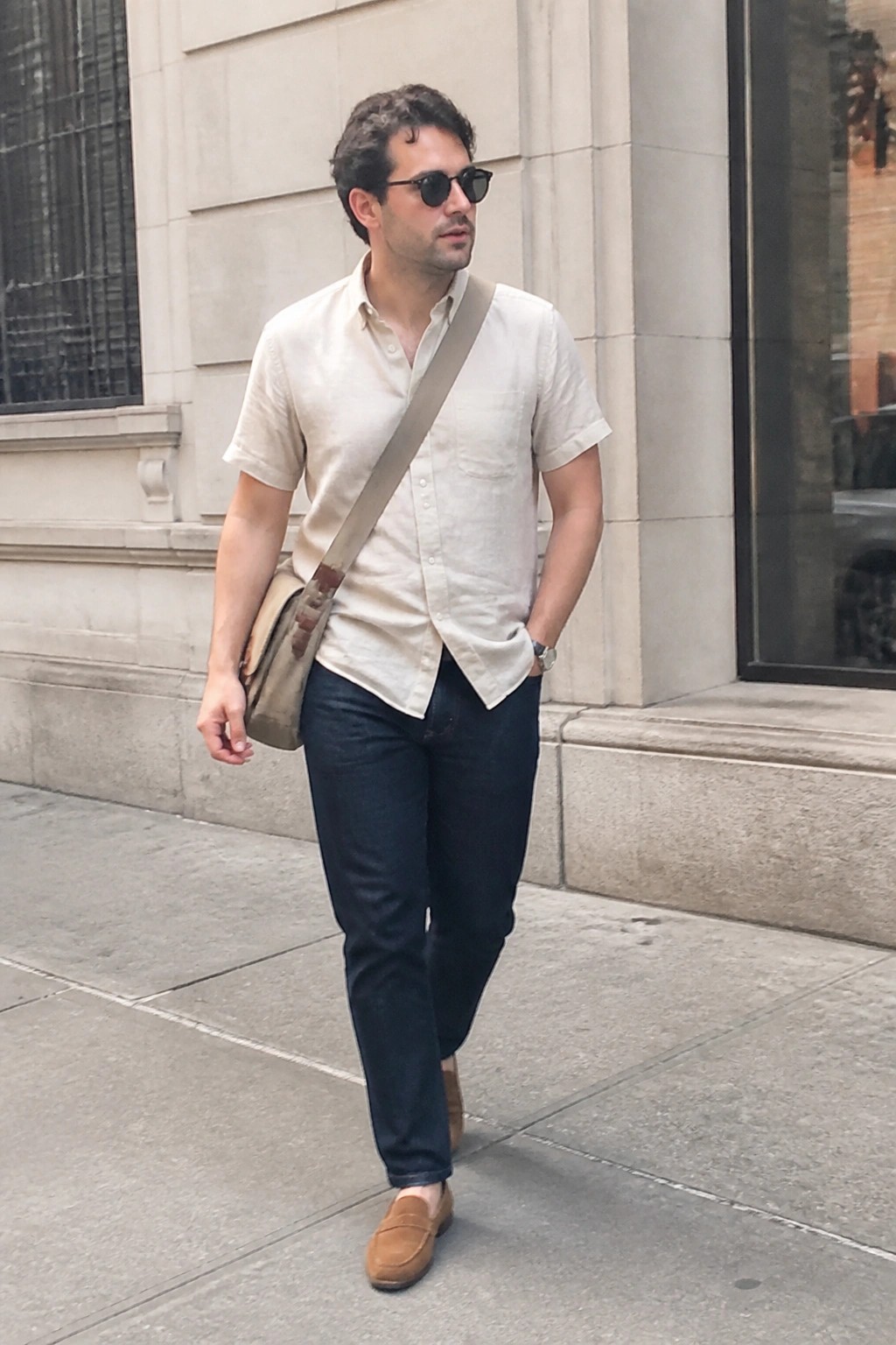 Man in beige shirt and dark jeans with crossbody bag on city sidewalk.