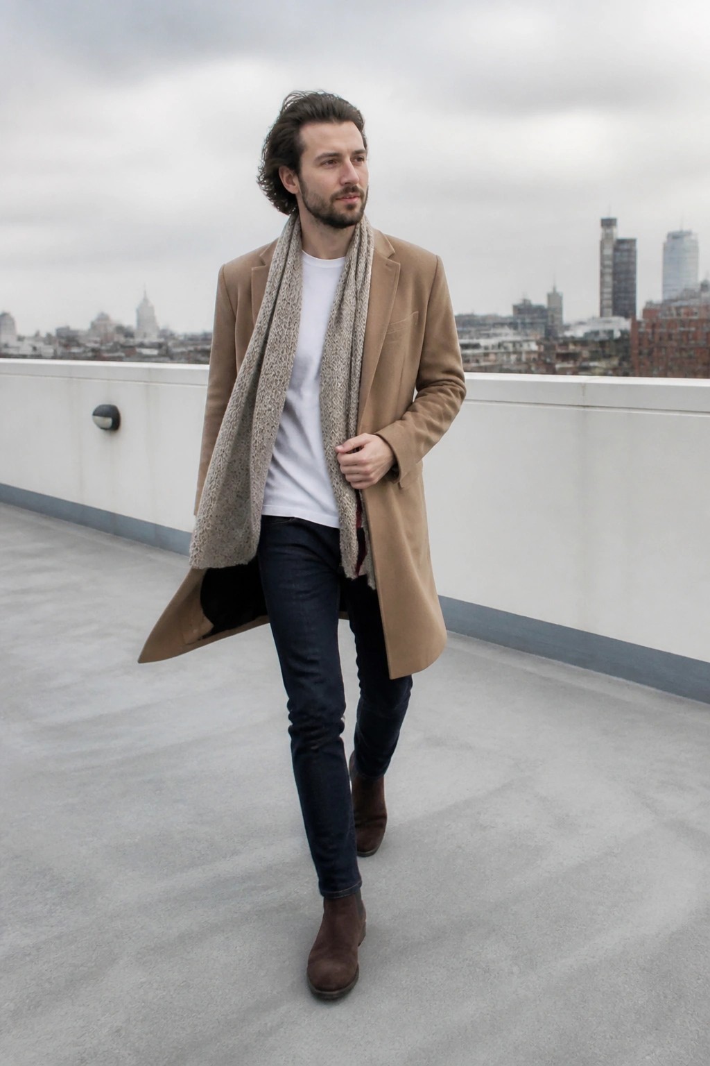 Man in a long camel coat over a white t-shirt with a patterned scarf, dark jeans, and brown suede boots standing on a rooftop.