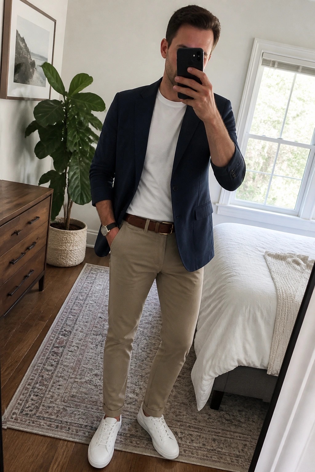 Man in navy blazer, white tee, and beige pants taking bedroom mirror selfie