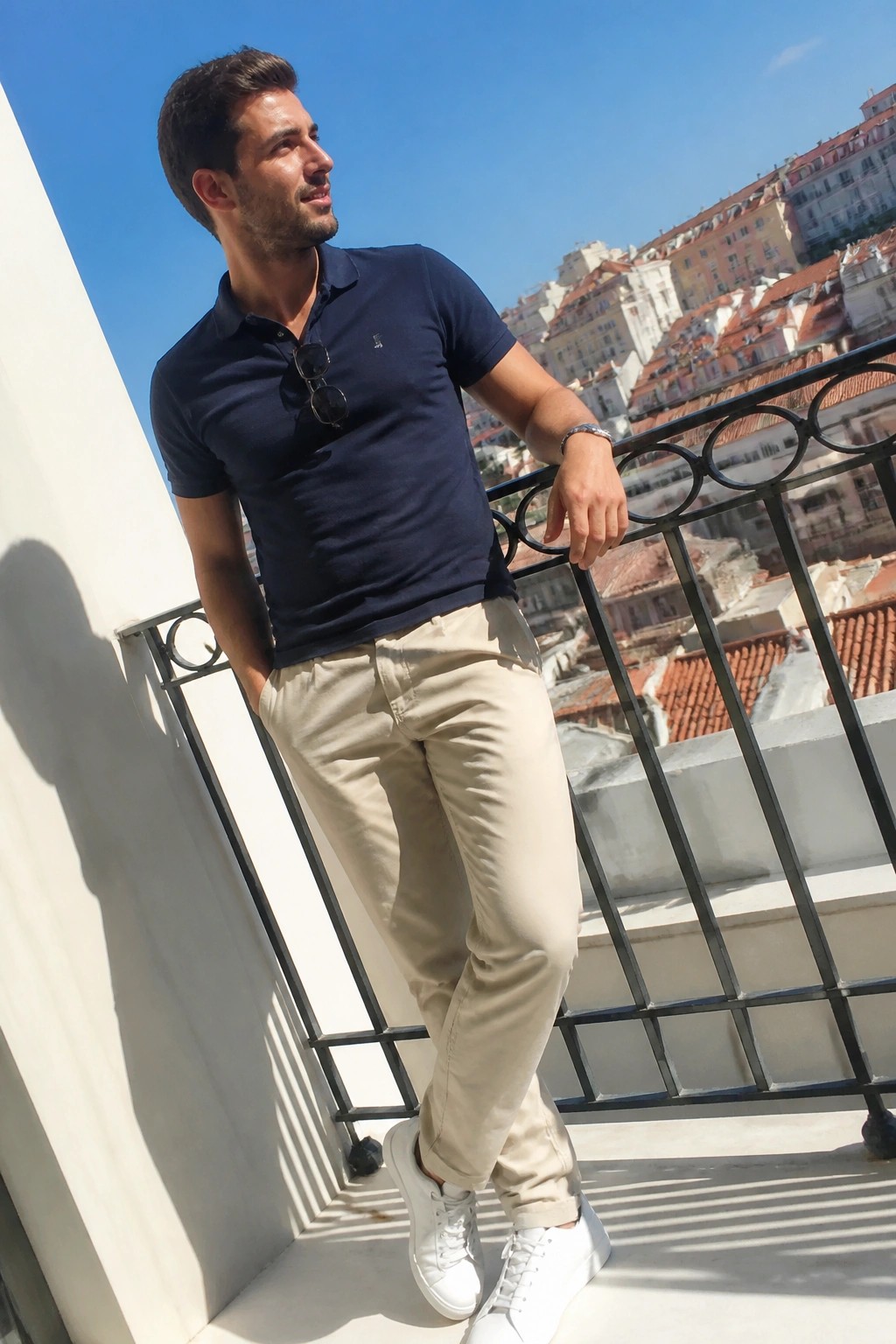 Man in navy polo shirt and beige pants leans on balcony railing above city.