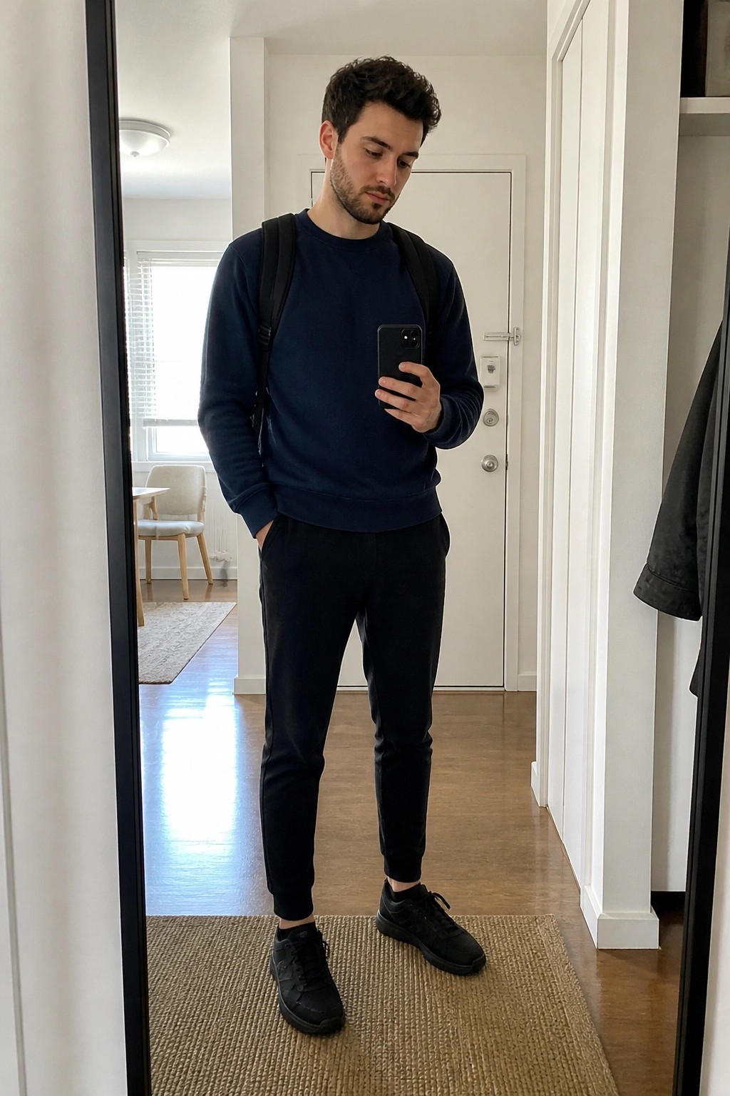 Man in navy sweatshirt and black pants taking mirror selfie with backpack