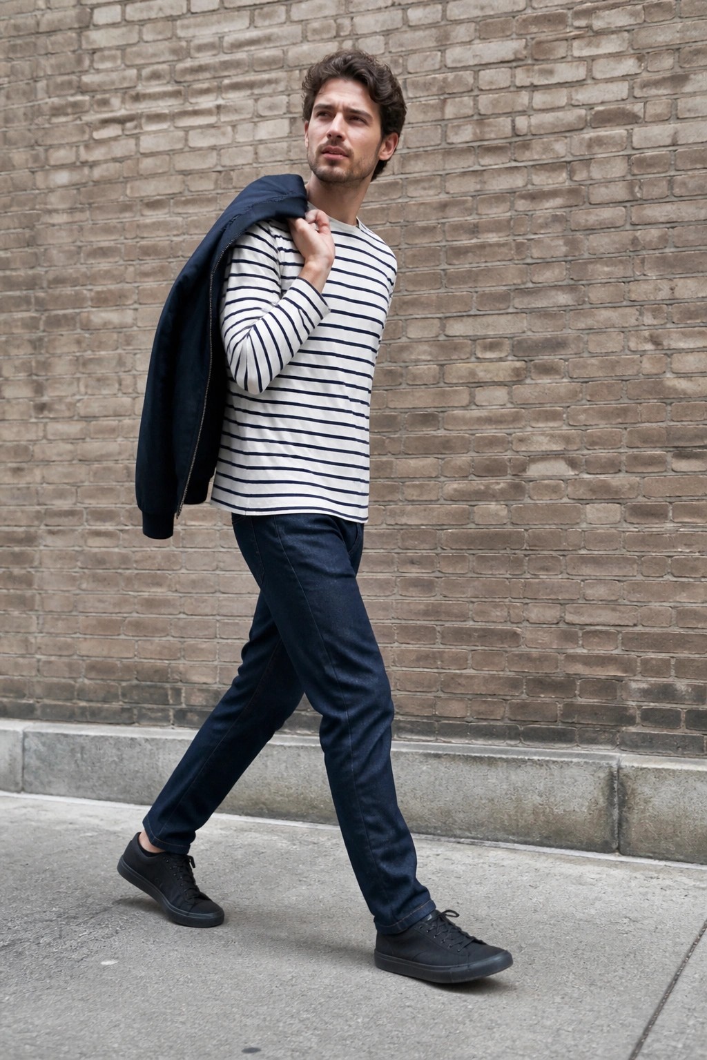 Man in striped shirt and jeans carrying navy jacket over shoulder by brick wall.