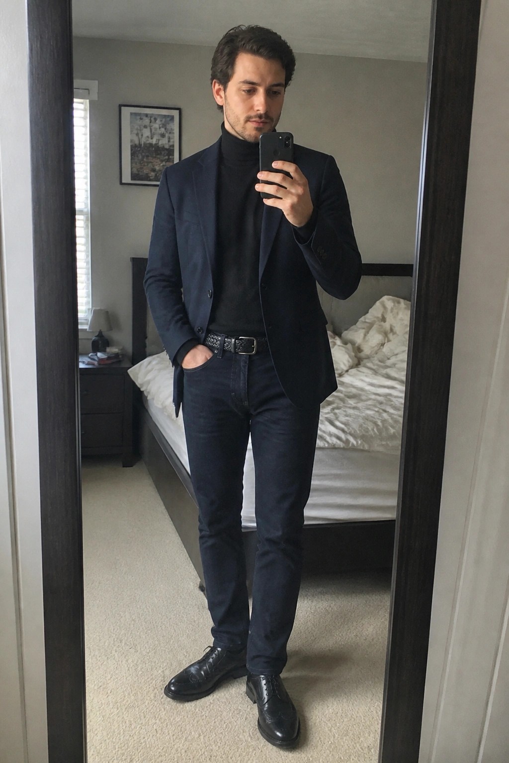 Man wearing navy blazer over black turtleneck with dark jeans black woven belt and black brogue shoes taking a mirror selfie in a bedroom