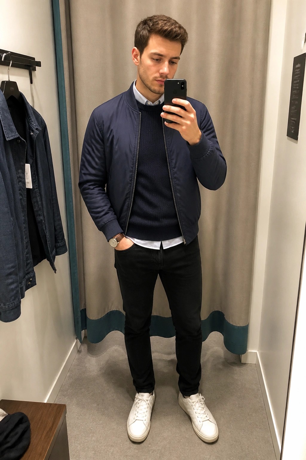 Man in navy bomber jacket, sweater, black jeans, white sneakers taking fitting room selfie