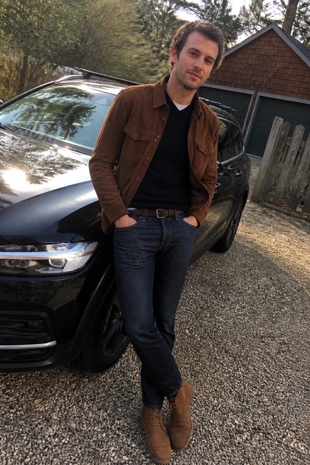 Man in brown suede jacket and jeans standing by black SUV on gravel