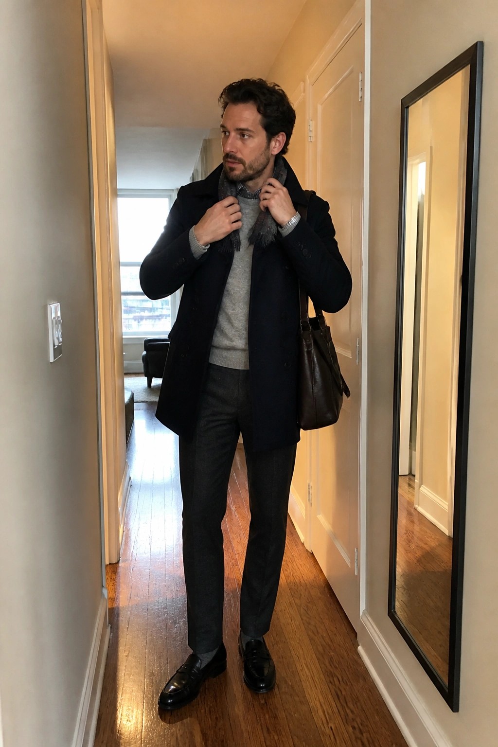 Man in navy overcoat over gray sweater adjusts scarf holding brown leather bag in hallway
