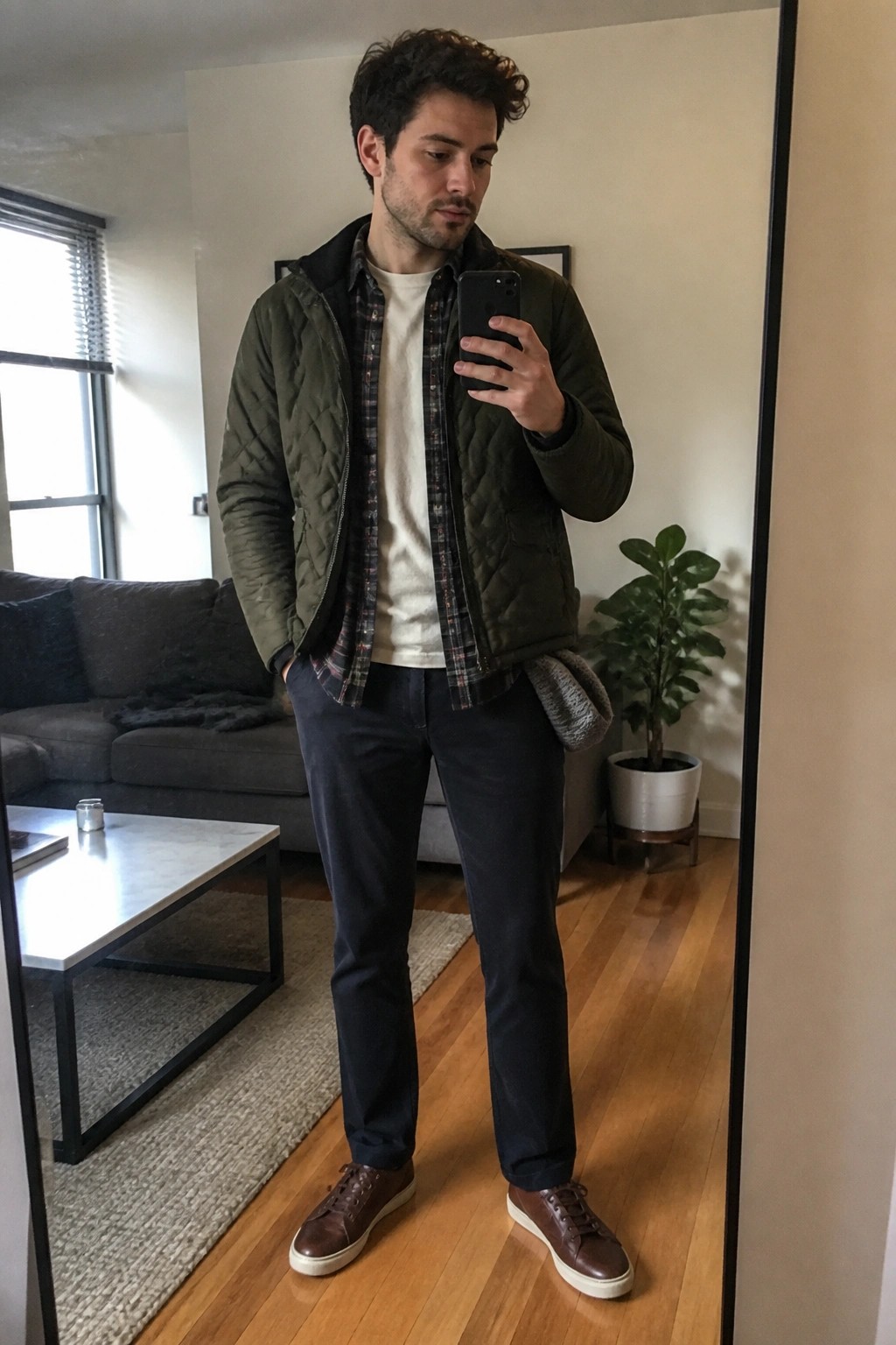 Man in green quilted jacket over plaid shirt taking mirror selfie in apartment