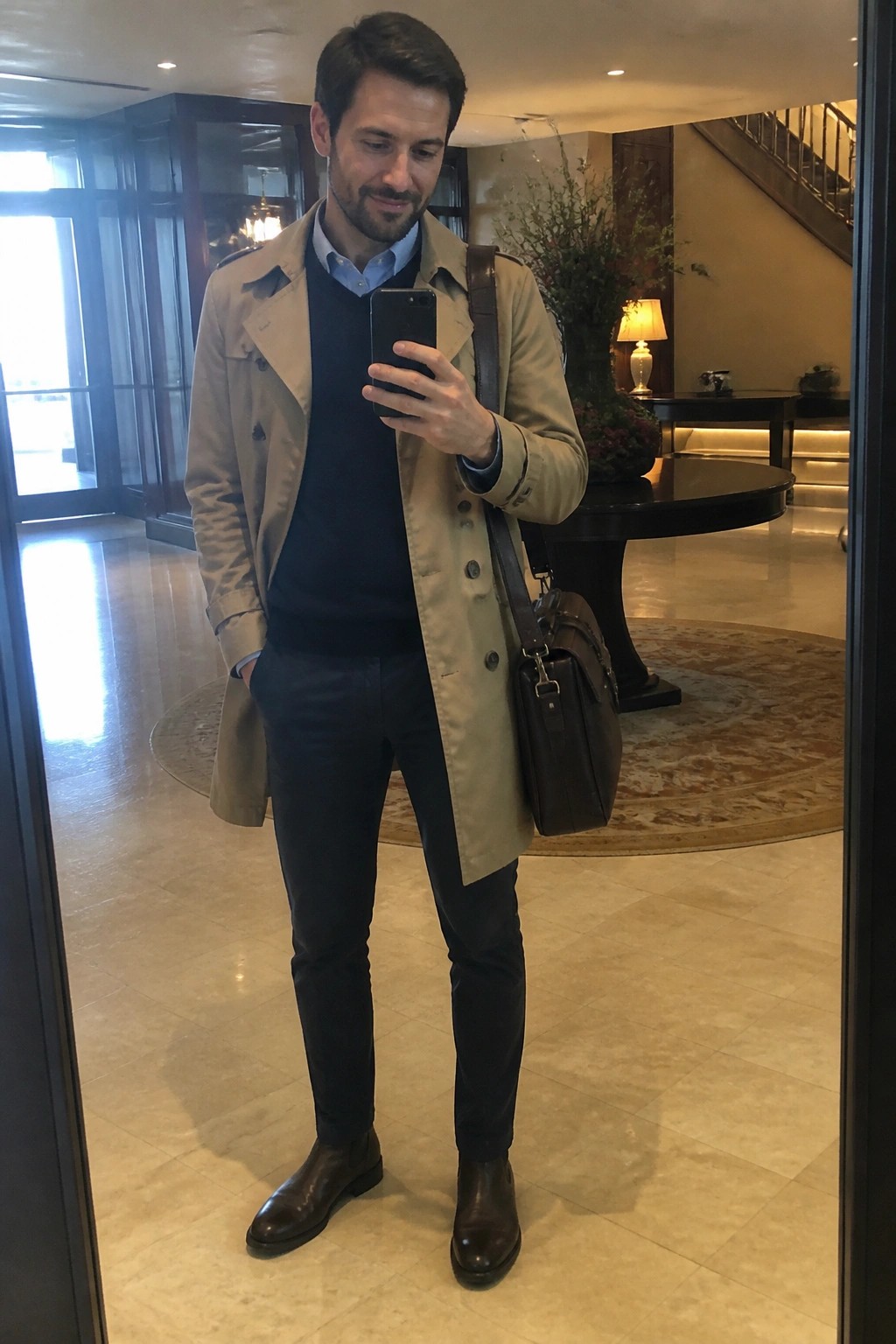 Man in beige trench coat taking mirror selfie with leather bag in elegant lobby