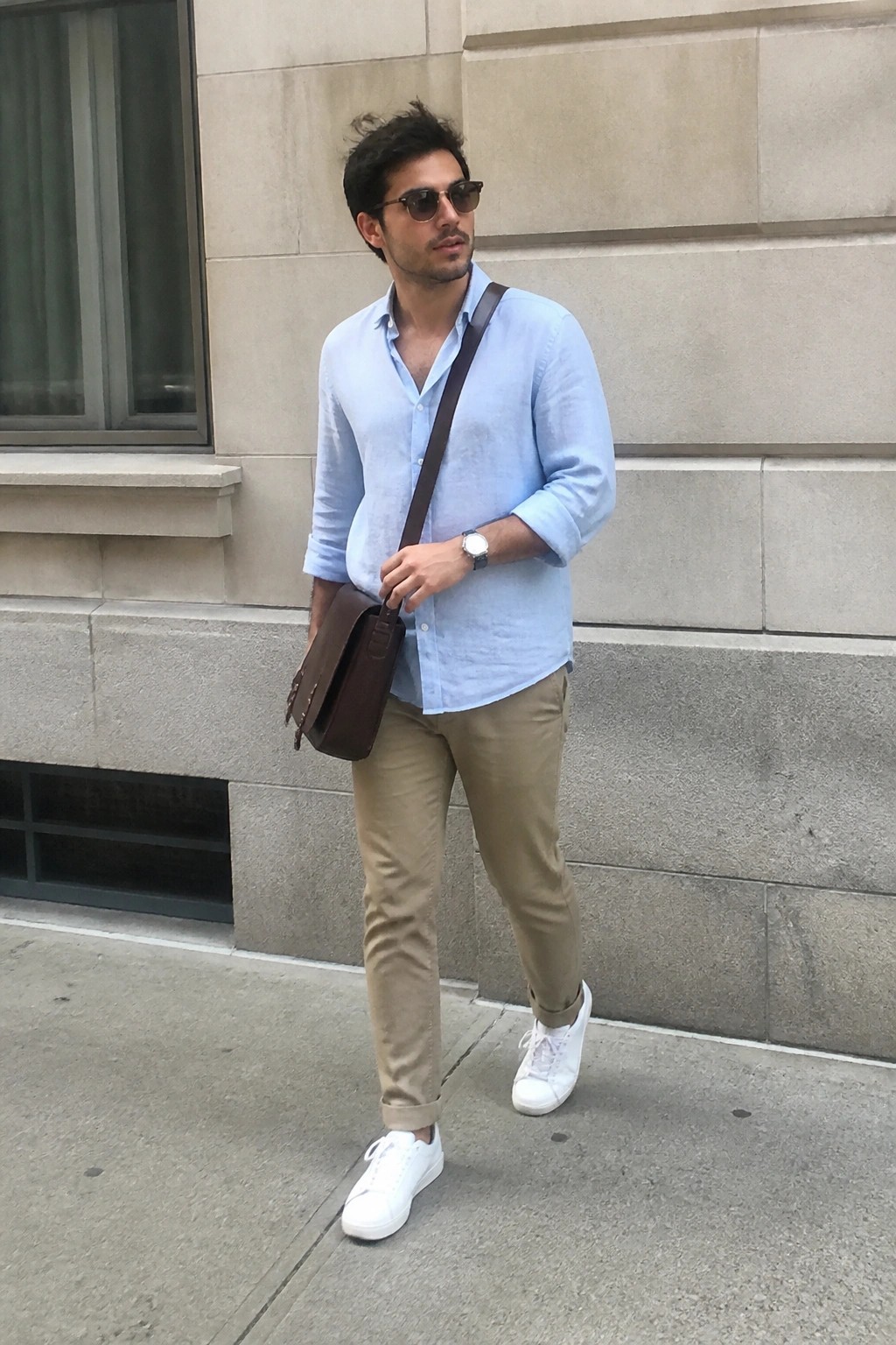 Man in light blue shirt, beige pants, white sneakers holding brown leather bag outdoors