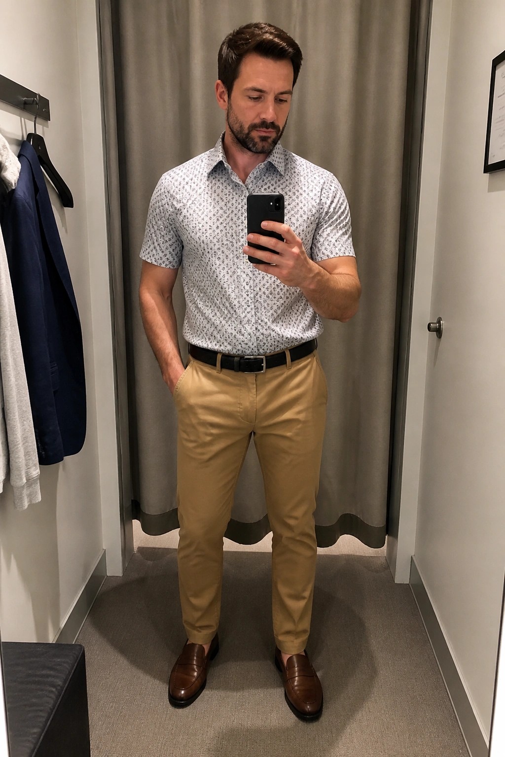 Man in patterned shirt and khaki pants taking mirror selfie in fitting room.