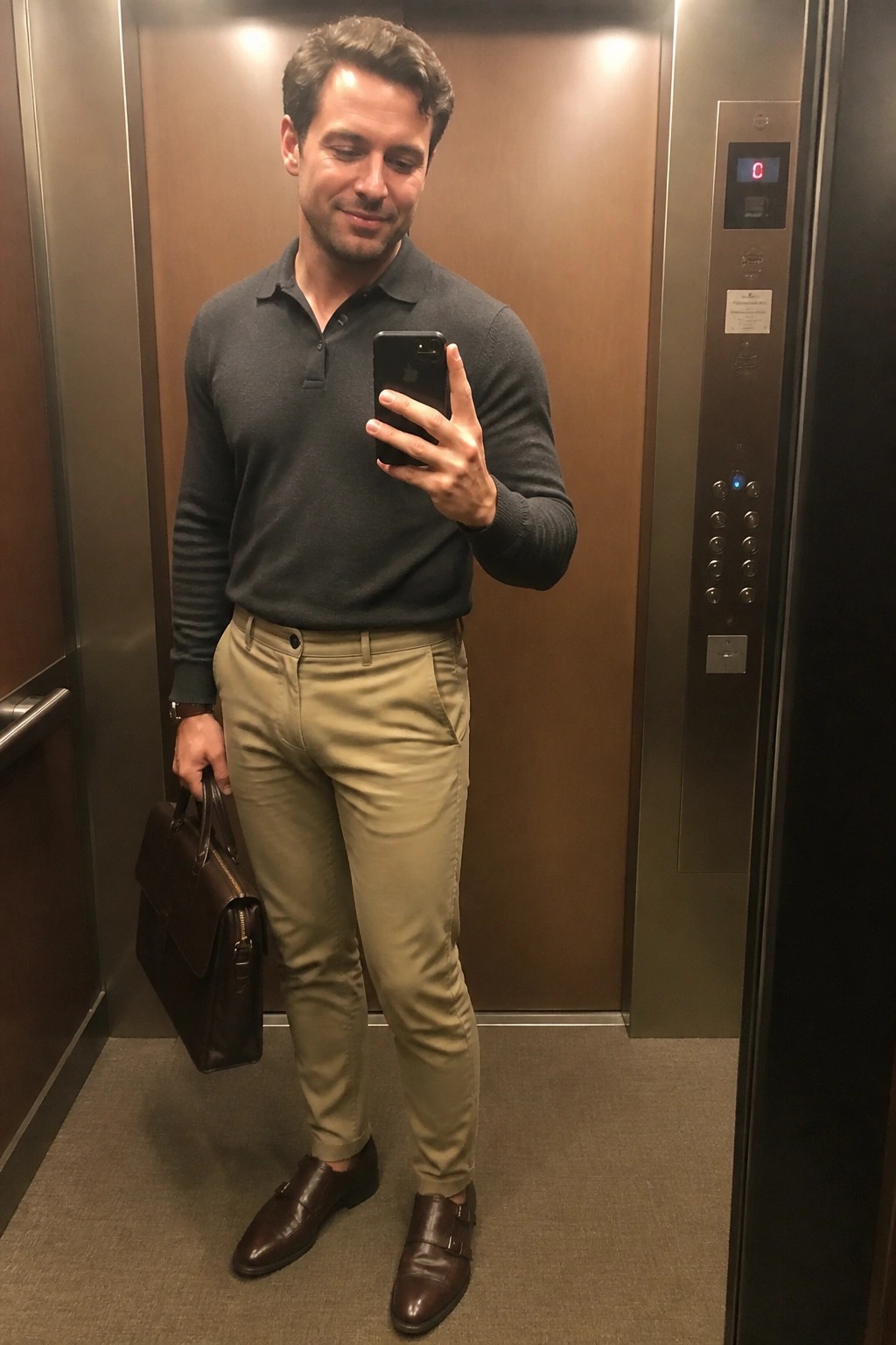 Man in gray polo shirt and khaki pants taking selfie in elevator with briefcase