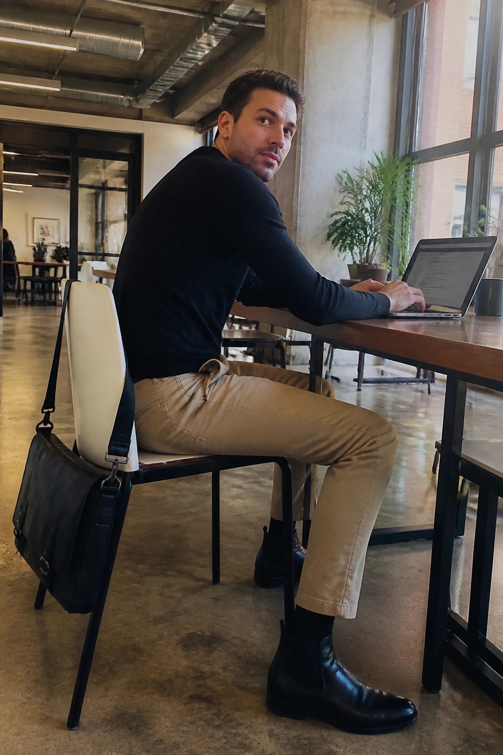 Man in black shirt and beige pants at wooden table with laptop and black bag