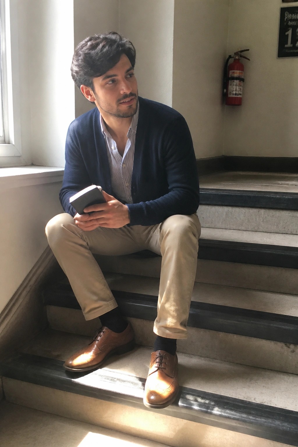 Man in navy cardigan sits on stairs holding book, wearing striped shirt and beige pants.