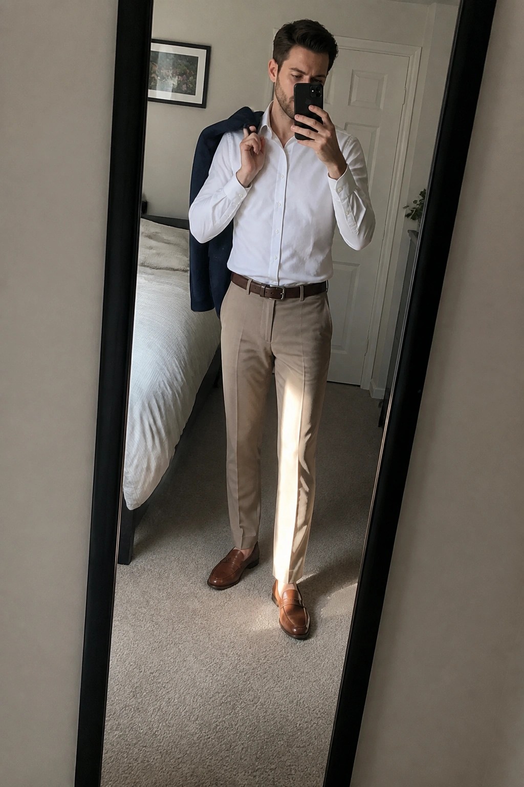 Man in white shirt and beige pants holding phone and navy blazer in mirror selfie