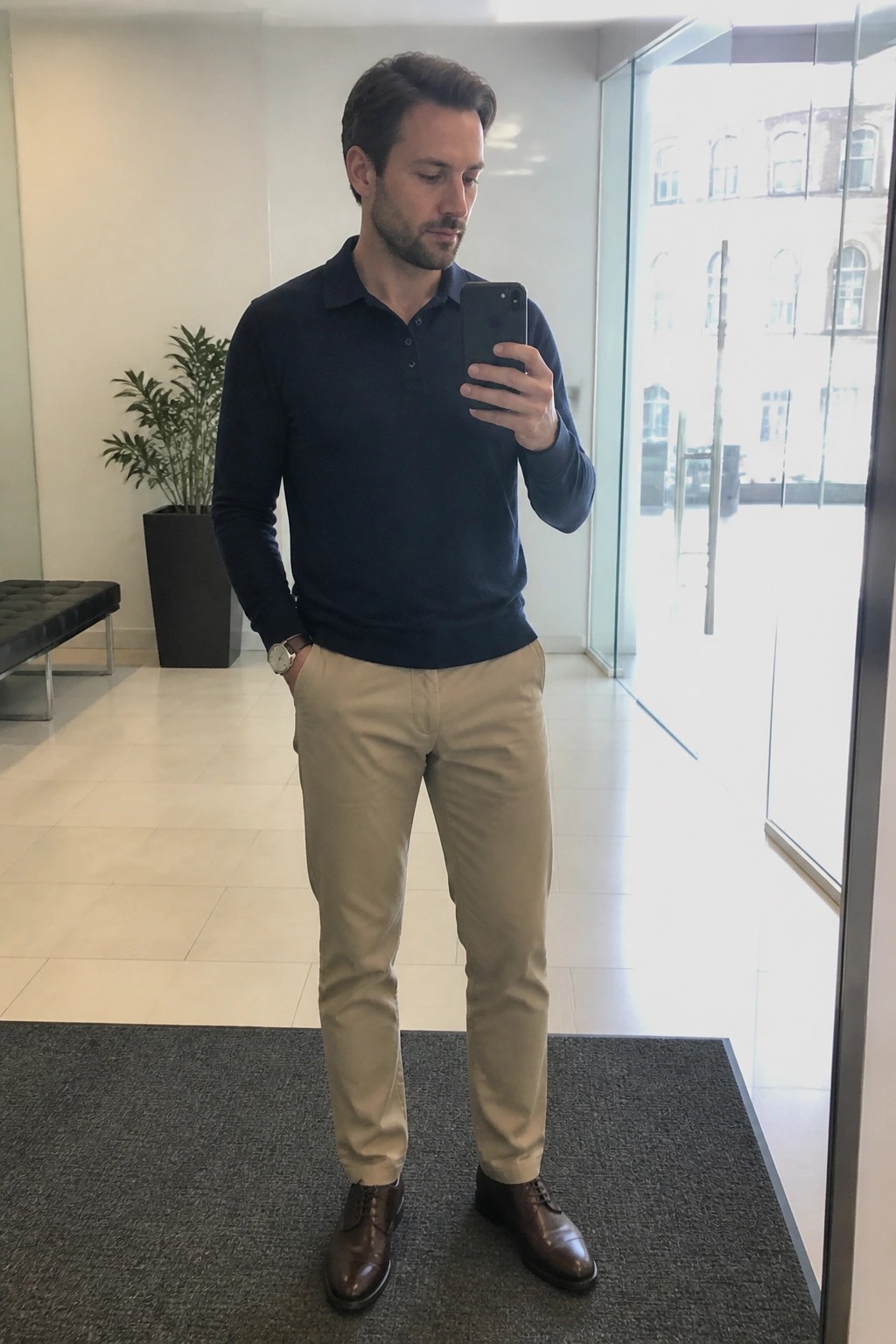 Man in navy polo shirt and khaki pants taking a mirror selfie indoors
