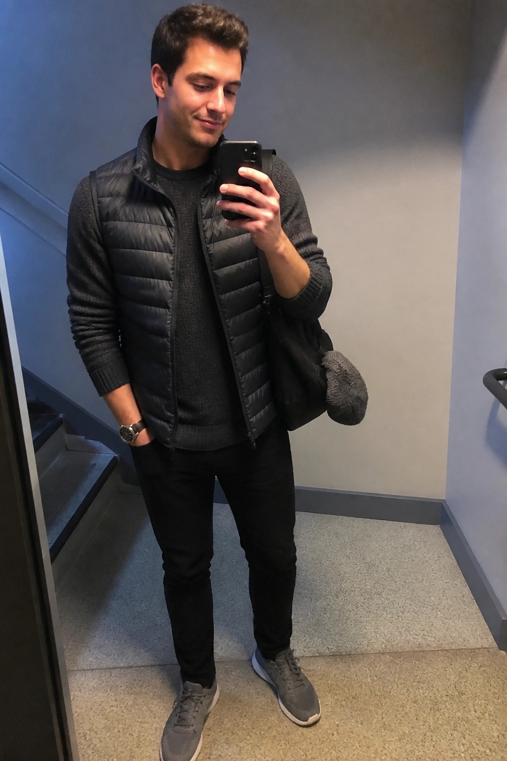 Man in black puffer vest and sweater taking phone mirror selfie with bag